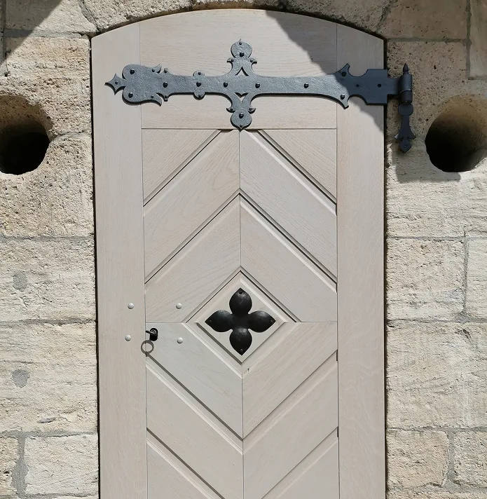 Helle Holztür mit Zierornament aus schwarzem Metall und Steinmauer mit zwei Öffnungen.