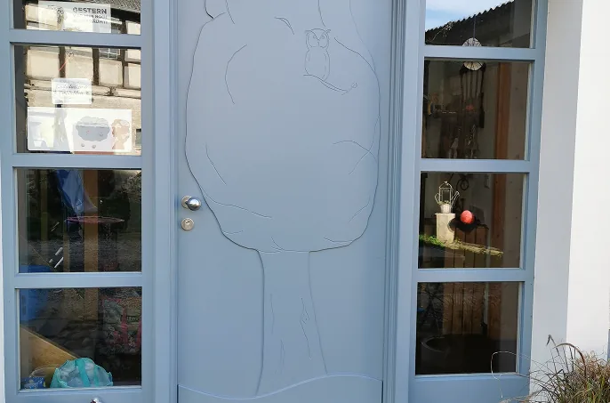 Grau gestrichene Holztür mit einem Baum und einer Eule als Gravur, flankiert von Fenstern.
