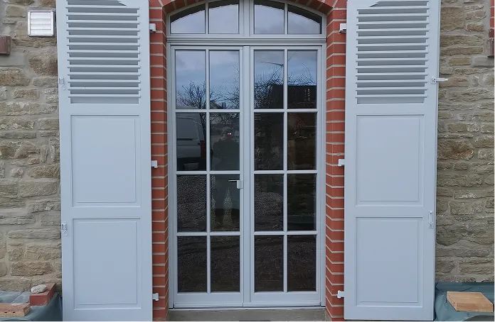Weiße Fensterläden neben zweiflügeliger Glastür mit rotem Backsteinrahmen an Steinmauer.