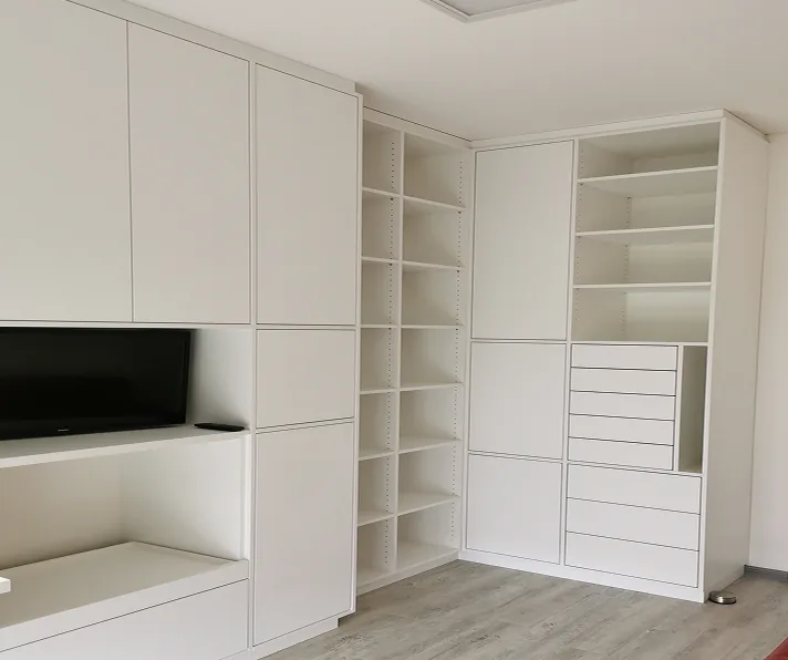 Moderne weiße Wohnzimmerschrankwand mit Regalen, Türen und Fernseher auf hellem Holzboden.