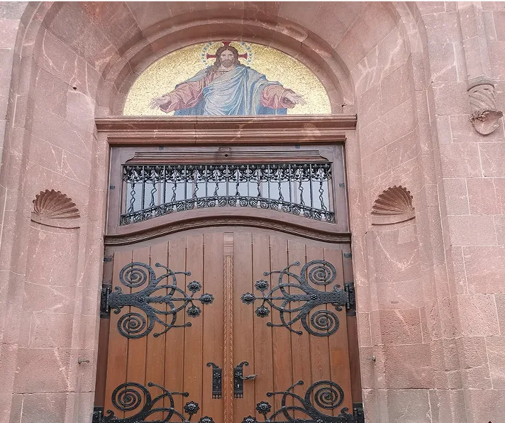 Holztür mit schmiedeeisernen Verzierungen und ein Mosaik von Jesus Christus darüber in einem Steinbogen.