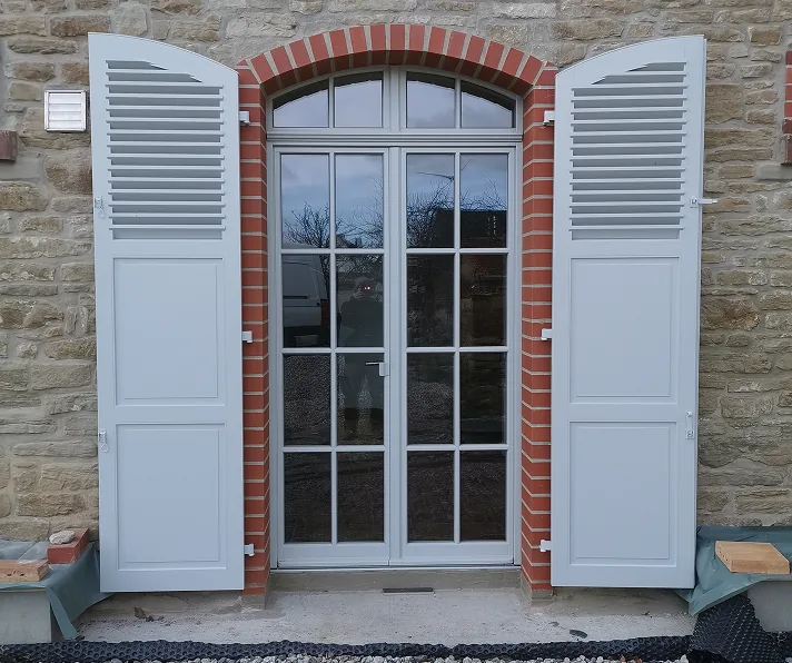Eine weiße Tür mit Glasfenstern und offenen weißen Fensterläden in einem Steinmauergebäude.