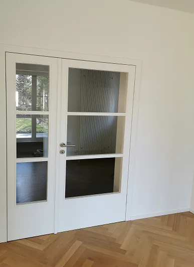 Weiße Innentür mit sechs Glasfenstern und Holzparkettboden in einem leeren Raum.